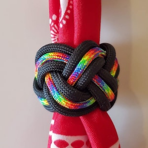 May include: A decorative black rope knot with a rainbow-colored center, wrapped around a red fabric with a white pattern. The knot is tightly woven, showcasing the vibrant rainbow hues against the black rope.