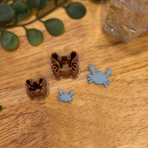 May include: Three cookie cutters in the shape of crabs. The largest cutter is brown and the two smaller cutters are blue.