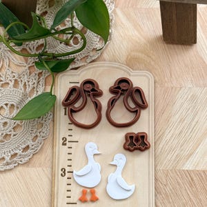 May include: Wooden duck-shaped cookie cutters and cut-out ducks. The cutters are brown, and the ducks are white with orange feet. The wooden base has an inch ruler along the side. The image is on a wooden surface.
