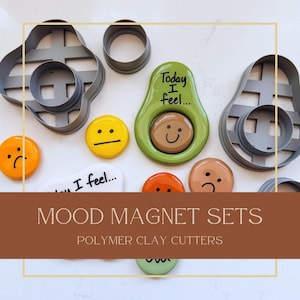 May include: A collection of gray polymer clay cutters alongside several circular mood magnets featuring various facial expressions. One green avocado-shaped magnet displays the text Today I feel... with a brown smiling face insert.