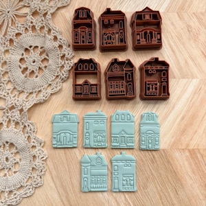 May include: A collection of house-shaped cookie cutters in two colors: brown and light blue. The cutters feature detailed architectural designs, including windows, doors, and rooflines. The cutters are arranged on a wooden surface.