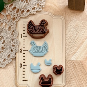 May include: A set of brown and blue chicken-shaped cookie cutters on a wooden measuring board. The cutters are in various sizes, with the largest measuring approximately 2 inches. The board has inch markings along the side.