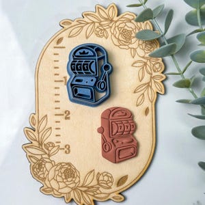 May include: Two wooden cookie cutters shaped like slot machines. The slot machine on the left is blue and the slot machine on the right is red. The cookie cutters are on a wooden background with a floral design.
