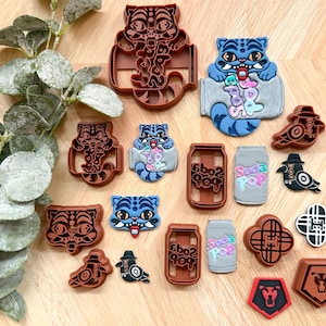 May include: A collection of colorful, decorative shoe charms and cookie cutters. The designs include a tiger, a soda can with the text "Soda Pop", and a bird wearing a hat. The color palette includes brown, blue, black, and red.