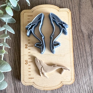 May include: A set of three whale-shaped cookie cutters on a wooden board with a ruler. The two larger cutters are blue and the smaller cutter is brown.