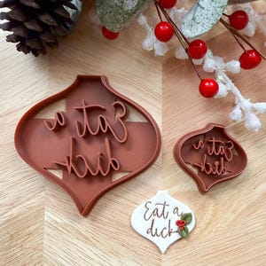May include: Brown plastic ornament-shaped cookie cutters with the words "o taz haib" and a white ornament with the words "Eat a duck" and holly. Festive holiday baking tools.