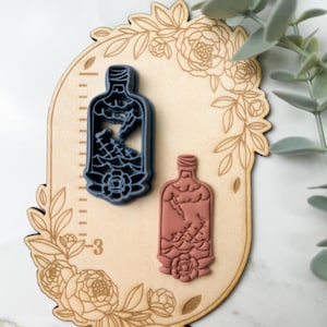 May include: Two cookie cutters shaped like bottles with a floral design on a wooden background. The bottle on the left is blue and the bottle on the right is pink.