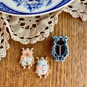 May include: Two floral patterned cookie cutters in the shape of a beetle, and a blue plastic beetle cookie cutter. The cookie cutters are on a wooden surface.