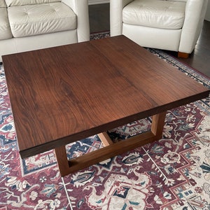 May include: A square wooden coffee table with a dark brown wood grain finish. The table has a simple, modern design with a rectangular base.