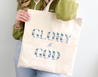 Può includere: Una borsa tote in tela bianca con le parole "Glory to God" stampate in lettere floreali blu.