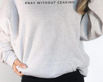 Può includere: Una felpa con cappuccio grigio chiaro con la scritta "PRAY WITHOUT CEASING" stampata in nero. La felpa con cappuccio ha un cordino e una tasca frontale. La persona indossa leggings neri.