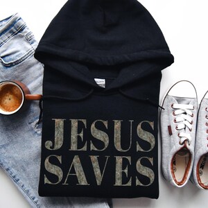 Puede incluir: Sudadera con capucha negra con el texto "JESUS SAVES" impreso en una fuente plateada desgastada. La sudadera se combina con jeans azul claro, una taza de caf&eacute; y zapatillas grises.