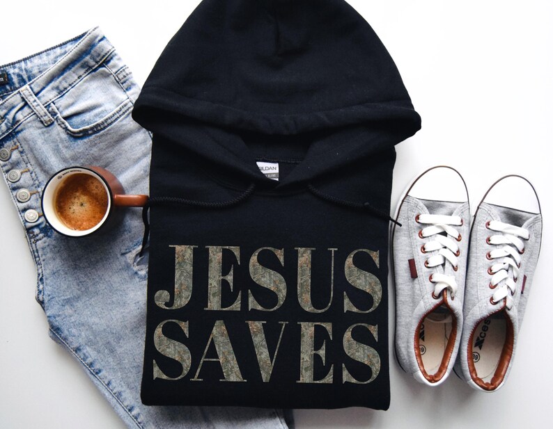 Puede incluir: Sudadera con capucha negra con el texto "JESUS SAVES" impreso en una fuente plateada desgastada. La sudadera se combina con jeans azul claro, una taza de caf&eacute; y zapatillas grises.