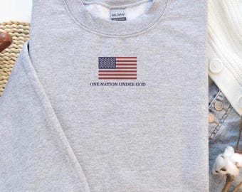 Può includere: Una felpa grigio chiaro con una bandiera americana ricamata in rosso, bianco e blu. Sotto la bandiera, il testo recita "ONE NATION UNDER GOD". La felpa è realizzata in un materiale morbido.