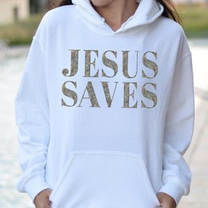 Puede incluir: Sudadera con capucha blanca con el texto "JESUS SAVES" impreso en un patr&oacute;n de camuflaje.