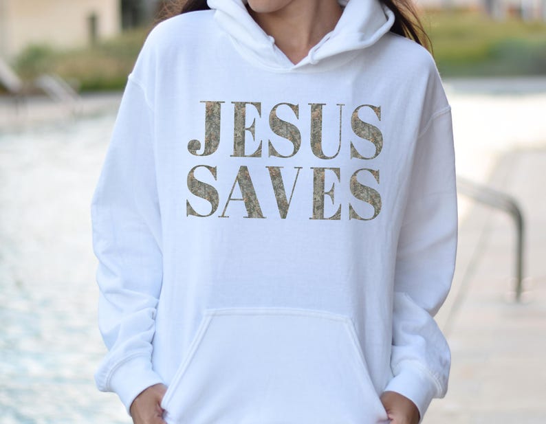 Puede incluir: Sudadera con capucha blanca con el texto "JESUS SAVES" impreso en un patr&oacute;n de camuflaje.