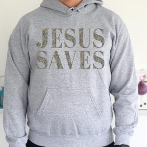 Puede incluir: Una sudadera con capucha gris con las palabras "JESUS SAVES" impresas en la parte delantera en un patr&oacute;n de camuflaje.