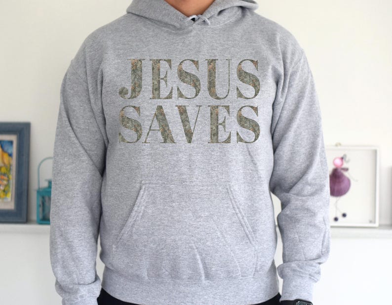 Puede incluir: Una sudadera con capucha gris con las palabras "JESUS SAVES" impresas en la parte delantera en un patr&oacute;n de camuflaje.