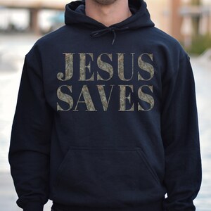 Puede incluir: Una sudadera con capucha azul oscuro con las palabras "JESUS SAVES" impresas en un patr&oacute;n de camuflaje en la parte delantera.