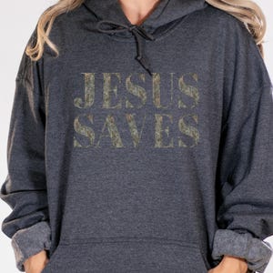 Puede incluir: Una sudadera con capucha gris oscuro con las palabras "JESUS SAVES" impresas en una fuente de estilo camuflaje desgastado.