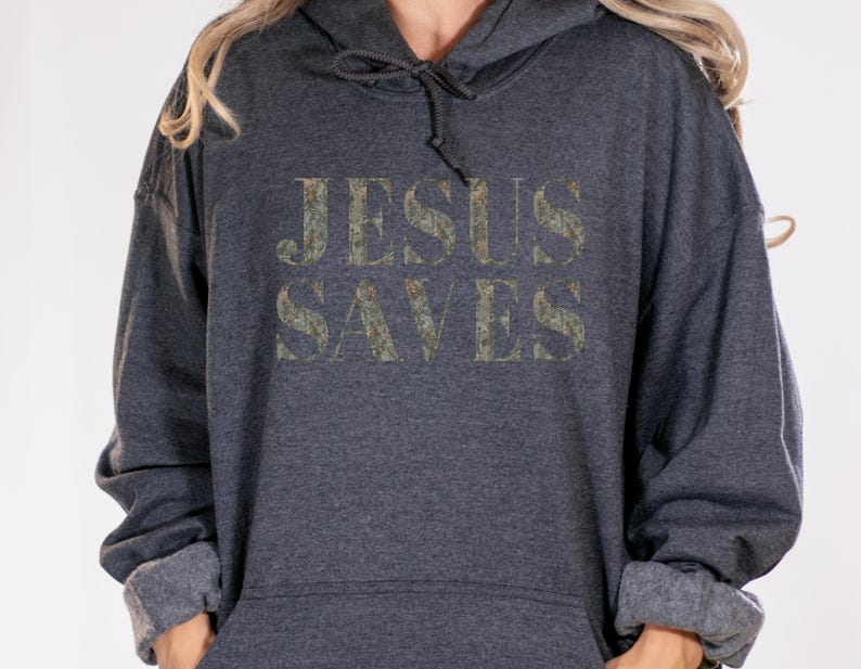 Puede incluir: Una sudadera con capucha gris oscuro con las palabras "JESUS SAVES" impresas en una fuente de estilo camuflaje desgastado.