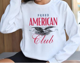 Può includere: Felpa bianca con un design grafico rosso, bianco e blu. La grafica presenta un'aquila con il testo "Proud American Club Est. 1776".