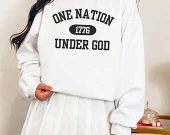 Puede incluir: Sudadera blanca con el texto negro "ONE NATION 1776 UNDER GOD". La sudadera es de cuello redondo. La imagen muestra a una persona que lleva la sudadera y una falda plisada blanca.