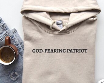 Può includere: Una felpa con cappuccio beige con la scritta "GOD-FEARING PATRIOT" in nero. La felpa con cappuccio è piegata e ha un cappuccio. Una tazza di caffè e un paio di jeans sono visibili sullo sfondo.