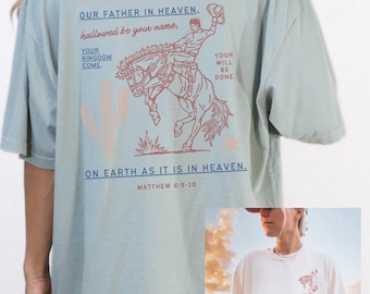 Può includere: Una t-shirt blu chiaro con una grafica di un cowboy che cavalca un cavallo che si impenna. La grafica è in rosso e marrone. Il testo "OUR FATHER IN HEAVEN. hallowed be your name, YOUR KINGDOM COME. YOUR WILL BE DONE. ON EARTH AS IT IS IN HEAVEN. MATTHEW 6:9-10" è stampato sulla t-shirt. La t-shirt è una "OUR FATHER" tee in stile western.