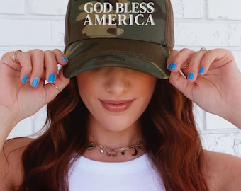Puede incluir: Gorra de camionero de camuflaje con el texto "GOD BLESS AMERICA" en blanco. La gorra tiene una parte trasera de malla y una visera curva. Una persona sostiene la gorra.