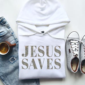 Puede incluir: Sudadera con capucha blanca con el texto "JESUS SAVES" impreso en una fuente gris desgastada. La sudadera est&aacute; colocada sobre una superficie blanca con un par de zapatillas de lona grises y un par de pantalones vaqueros azules.
