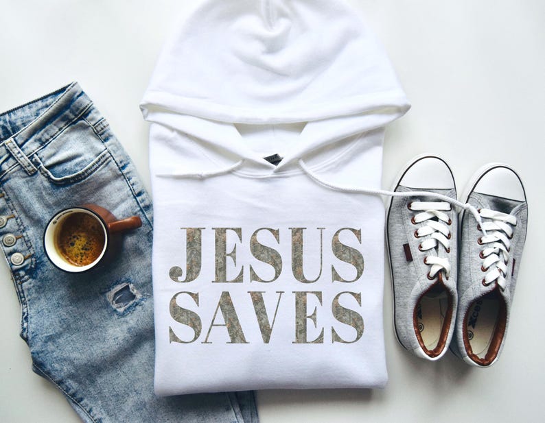 Puede incluir: Sudadera con capucha blanca con el texto "JESUS SAVES" impreso en una fuente gris desgastada. La sudadera est&aacute; colocada sobre una superficie blanca con un par de zapatillas de lona grises y un par de pantalones vaqueros azules.