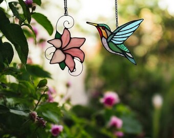 May include: Two stained glass suncatchers hanging from a window. One is a pink flower and the other is a green and blue hummingbird.