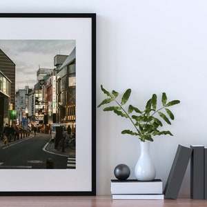 May include: A framed black and white photograph of a street in Japan at dusk. The street is lined with buildings and there are people walking on the sidewalk. The photo is hanging on a white wall with a vase of green plants and a stack of books on a table below it.