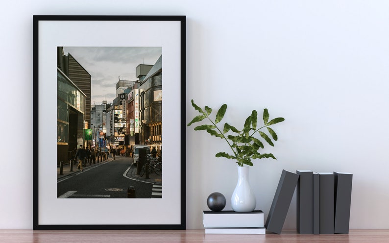 May include: A framed black and white photograph of a street in Japan at dusk. The street is lined with buildings and there are people walking on the sidewalk. The photo is hanging on a white wall with a vase of green plants and a stack of books on a table below it.