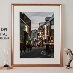 May include: A framed photograph of a street in Japan at dusk. The street is lined with buildings with neon signs. People are walking on the street.