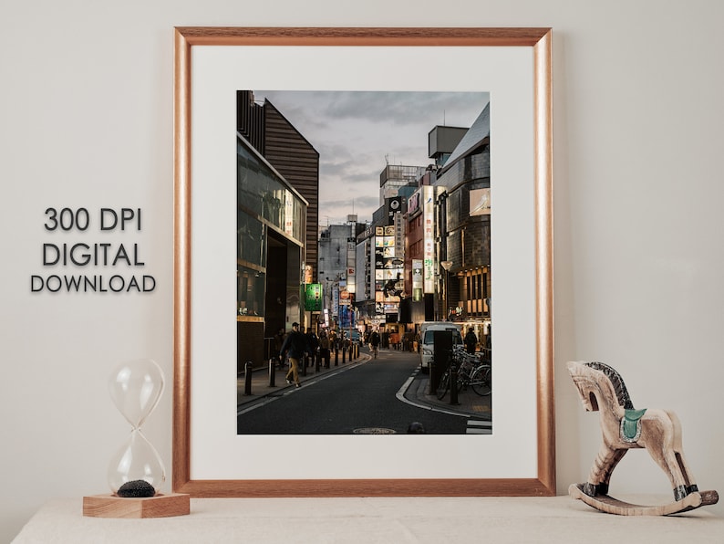 May include: A framed photograph of a street in Japan at dusk. The street is lined with buildings with neon signs. People are walking on the street.