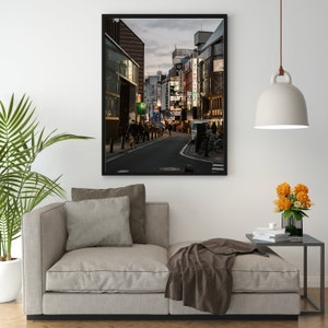 May include: A framed photograph of a street scene in Japan. The street is lined with buildings, some of which have neon signs. People are walking down the street, and there are cars parked on the side of the road.