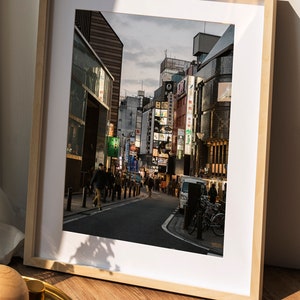 May include: A framed photograph of a street in Japan with buildings and neon signs. The street is lined with people walking and there are cars parked on the side of the road.