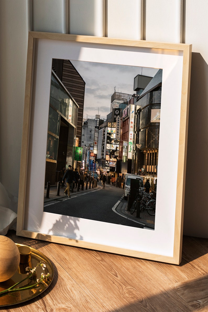 May include: A framed photograph of a street in Japan with buildings and neon signs. The street is lined with people walking and there are cars parked on the side of the road.