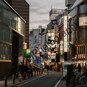 May include: A street scene in a city with tall buildings and many signs in Japanese. The street is lined with people walking and there are a few cars and bicycles parked on the side of the road.