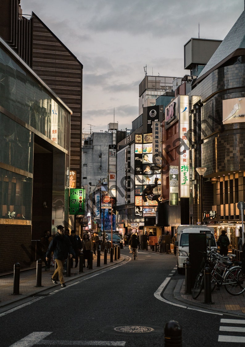 May include: A street scene in a city with tall buildings and many signs in Japanese. The street is lined with people walking and there are a few cars and bicycles parked on the side of the road.