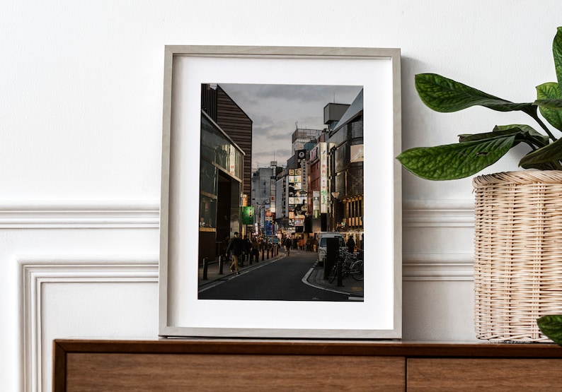 May include: A framed photograph of a street in Japan at dusk. The street is lined with buildings and shops, and there are people walking on the sidewalk. The buildings are lit up with neon signs, and the sky is a soft blue.