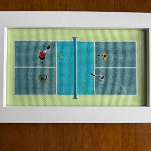Op de afbeelding: Kruissteekkunstwerk van een pickleballveld, wit omlijst. Het veld is turquoise en groen, met spelers in actie. Een gele bal bevindt zich in het midden. Het frame is rechthoekig en ligt op een houten oppervlak.