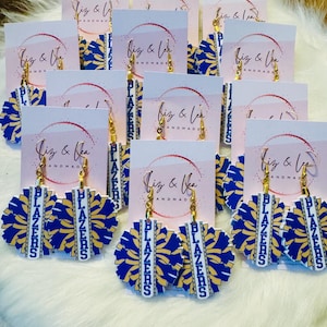 May include: A set of blue and gold pom-pom earrings with the word "BLAZERS" written on the dangle charms. The earrings are handmade and come with a white card that says "Biz & Lea Handmade".