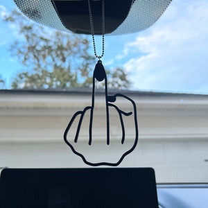 Op de afbeelding: Een zwarte autoluchtverfrisser in de vorm van een middelvinger die aan een achteruitkijkspiegel hangt.