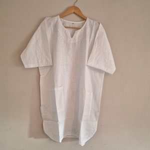 May include: White tunic shirt with short sleeves, a V-neck, and two front pockets. The shirt features embroidered detailing down the front and is hanging on a wooden hanger against a neutral wall.