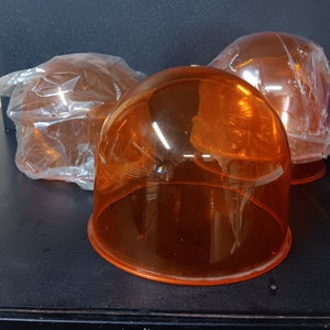 May include: Three orange-tinted, dome-shaped plastic covers, possibly for lights or equipment, are displayed on a dark surface. Two are wrapped in clear plastic, while one is unwrapped, showcasing its translucent quality. The items are likely for industrial or commercial use.