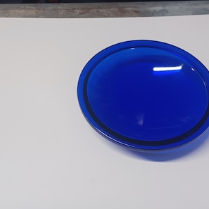 May include: A cobalt blue glass plate with a slightly raised rim. The plate is round and has a smooth, glossy finish.