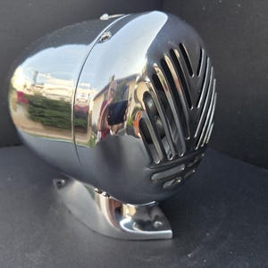 May include: A chrome-plated, retro-style speaker with a rounded body and a slotted front panel. The speaker sits on a matching base, reflecting the surrounding environment. The design evokes a vintage aesthetic.